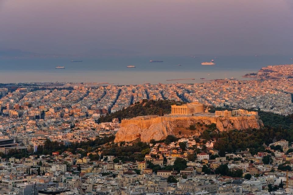 Ruinas antiguas en una colina brillan al atardecer, con vistas a un denso paisaje urbano que se extiende hasta el mar.