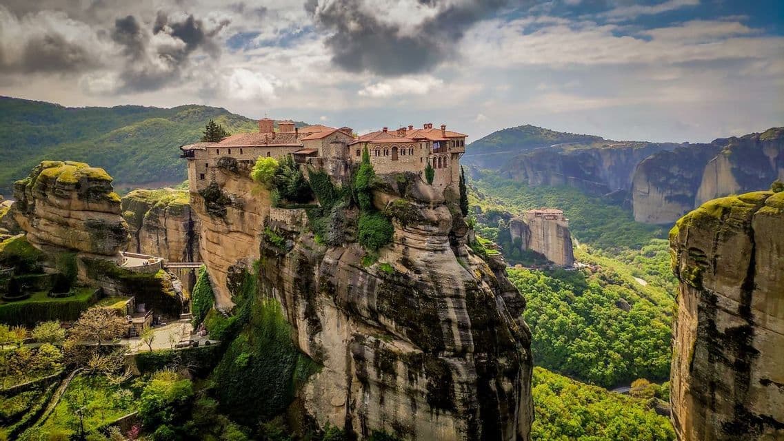 A stone monastery with a red-tiled roof sits atop a tall, sheer rock pillar, overlooking a vast, green, mountainous valley.