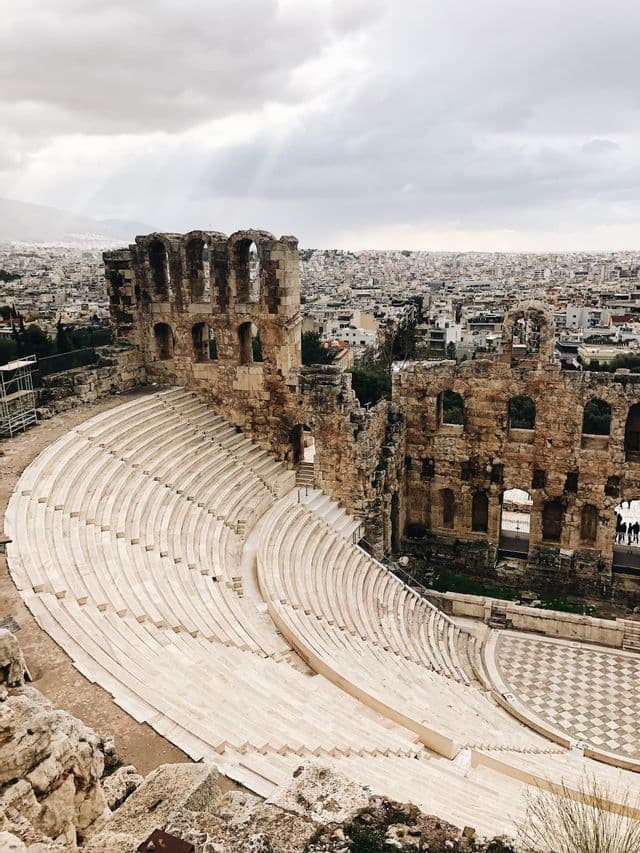 An ancient stone amphitheater with tiered seating overlooks a dense cityscape under a cloudy sky.