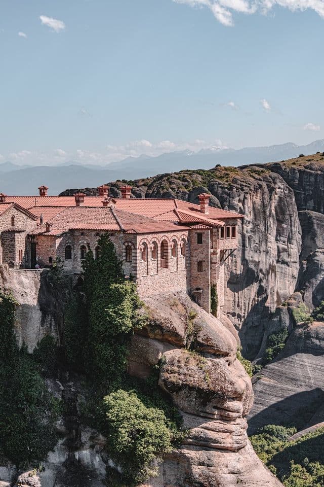 Ein steinernes Kloster mit rotem Ziegeldach, am Rande einer steilen Felsklippe gebaut, mit fernen Bergen im Hintergrund.