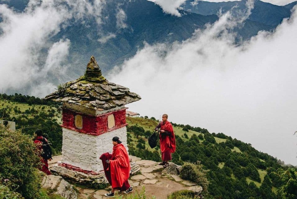 Due monaci in tuniche rosse si ergono accanto a una piccola stupa su un sentiero di montagna che domina nuvole e verdi colline.