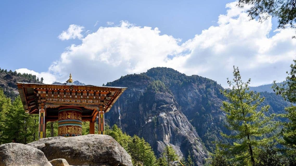 Una ruota di preghiera colorata in un padiglione di legno su una roccia, che si affaccia su una montagna boscosa con un monastero sulla parete della scogliera.