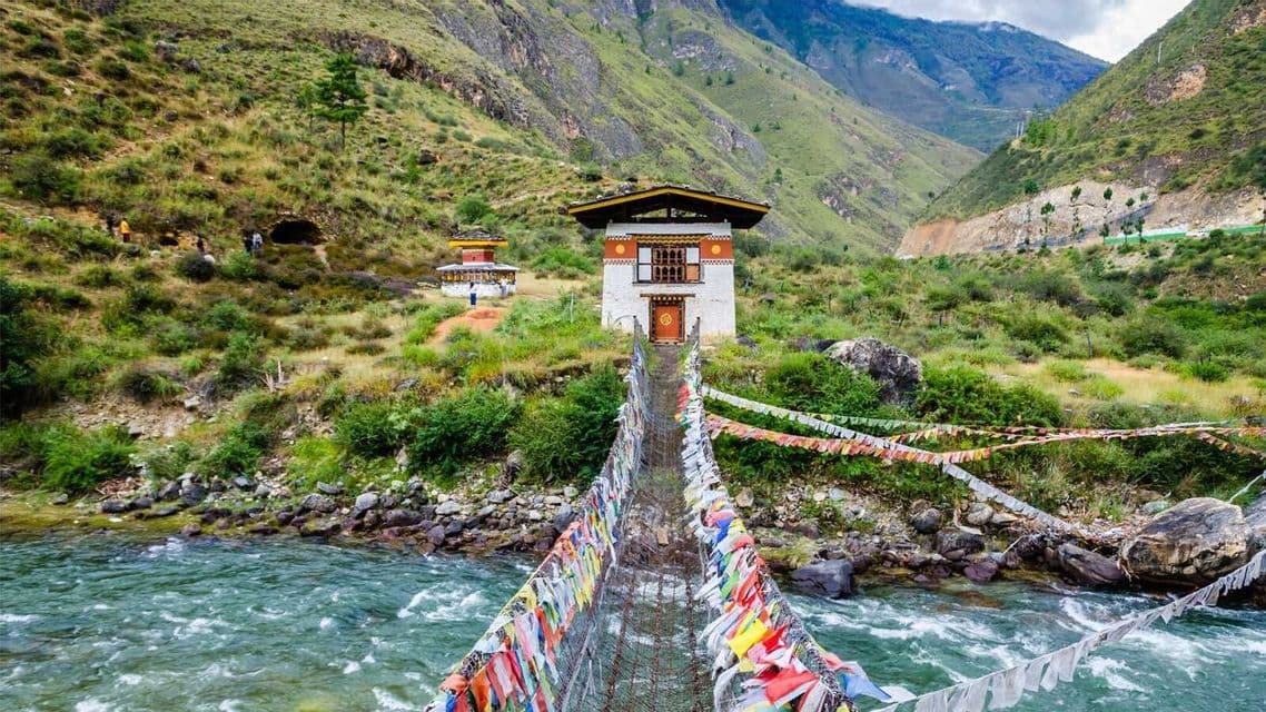Un ponte sospeso, adornato da colorate bandiere di preghiera, attraversa un ampio fiume e conduce a edifici tradizionali in una verde valle di montagna.