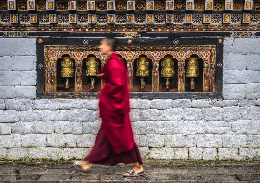 Un monaco in abiti rossi è immortalato con effetto mosso mentre cammina accanto a un muro di pietra con ruote di preghiera dorate.