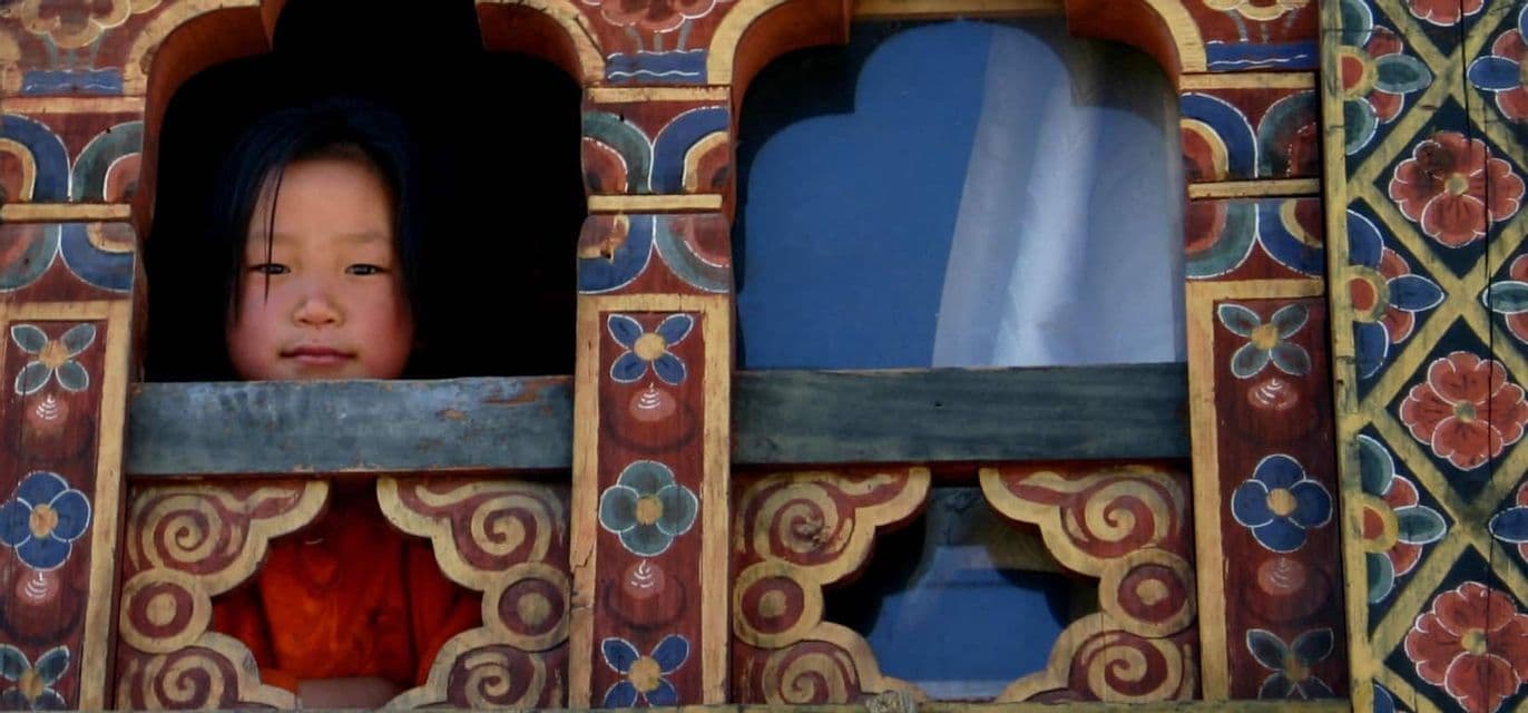 Un bambino con una maglietta arancione si affaccia da dietro una cornice di finestra in legno colorata e intagliata.