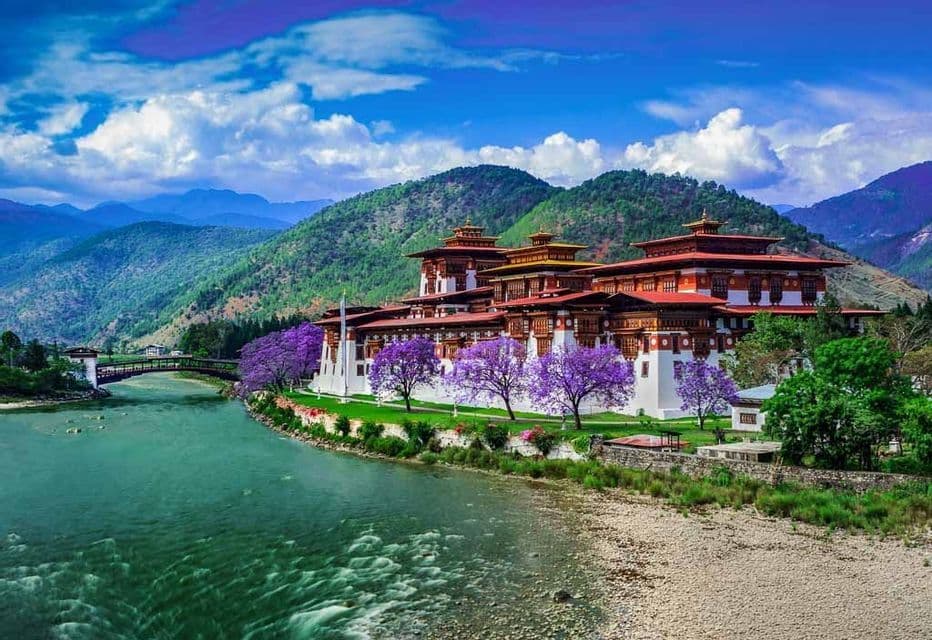Un grande monastero con tetti rossi e mura bianche sorge vicino a un fiume, con alberi fioriti viola e verdi colline alle sue spalle.