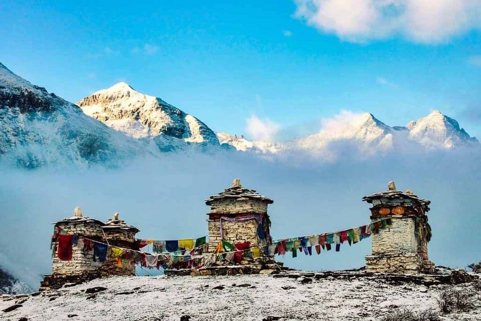 Bandiere di preghiera colorate appese tra due stupa di pietra su una collina innevata, con imponenti cime montuose che emergono dalle nuvole.