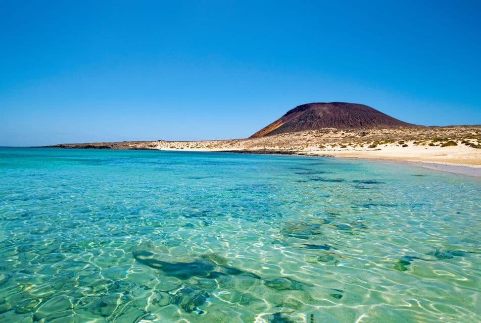 Acqua limpida e turchese lambisce una spiaggia sabbiosa di fronte a una grande collina vulcanica sotto un cielo azzurro senza nuvole.