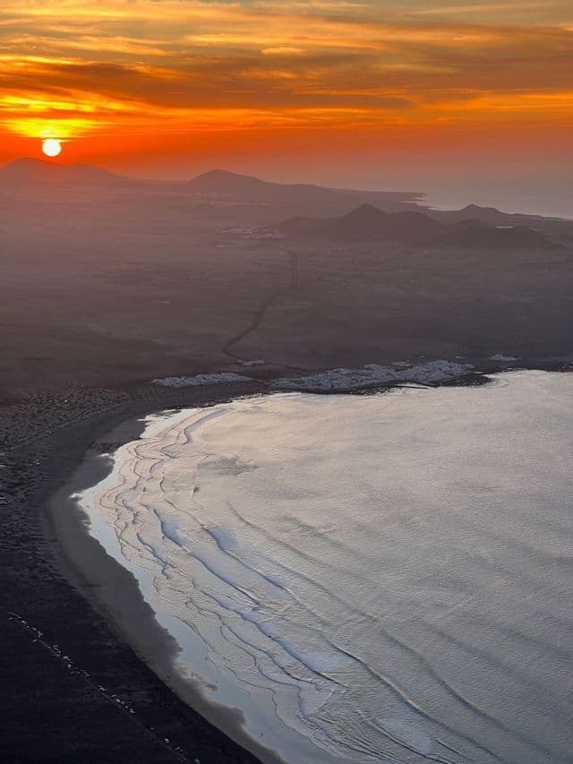 Veduta aerea di un vivace tramonto arancione su una costa montuosa con spiaggia di sabbia scura e onde dolci