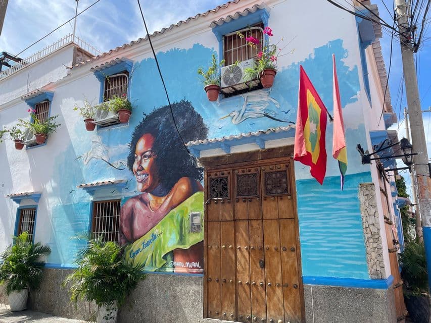 Un gran mural de una mujer sonriente pintado en la esquina de un edificio blanco y azul con una puerta de madera, banderas y macetas.