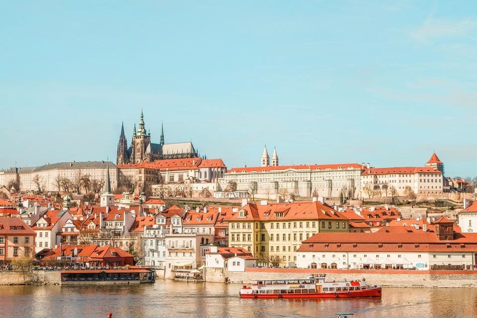 Una vista panorámica de una ciudad histórica con edificios de tejados rojos y un castillo en una colina, con un barco turístico en el río abajo.