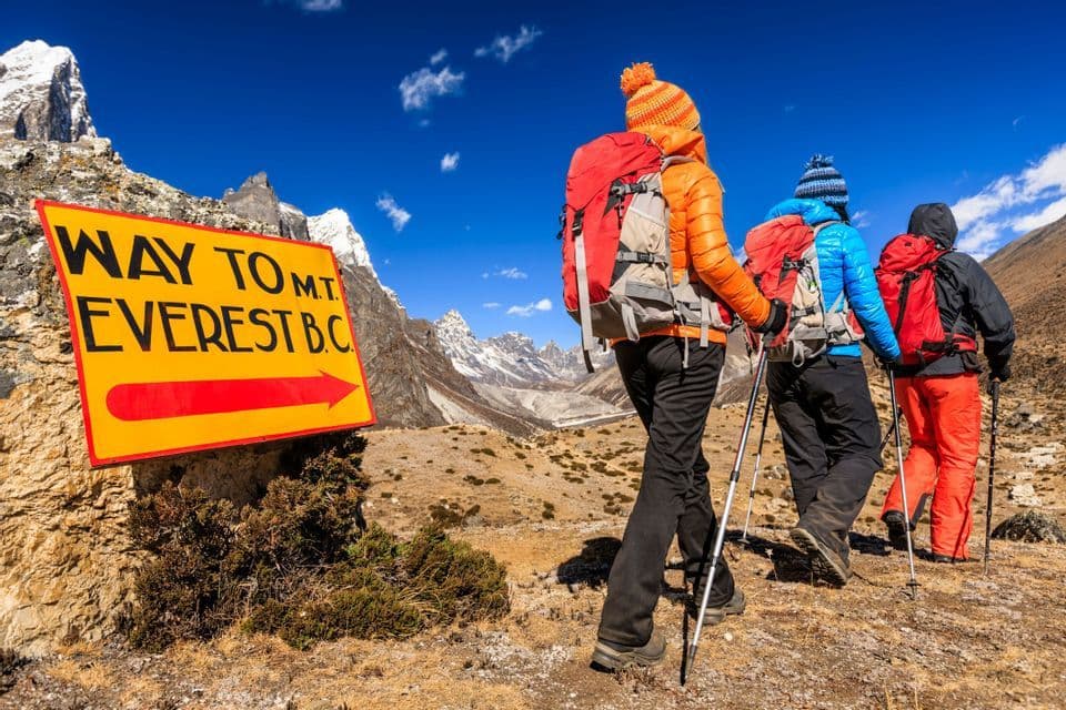 Un grupo WeRoad de tres excursionistas con mochilas y bastones camina junto a una señal direccional amarilla en un sendero de montaña.