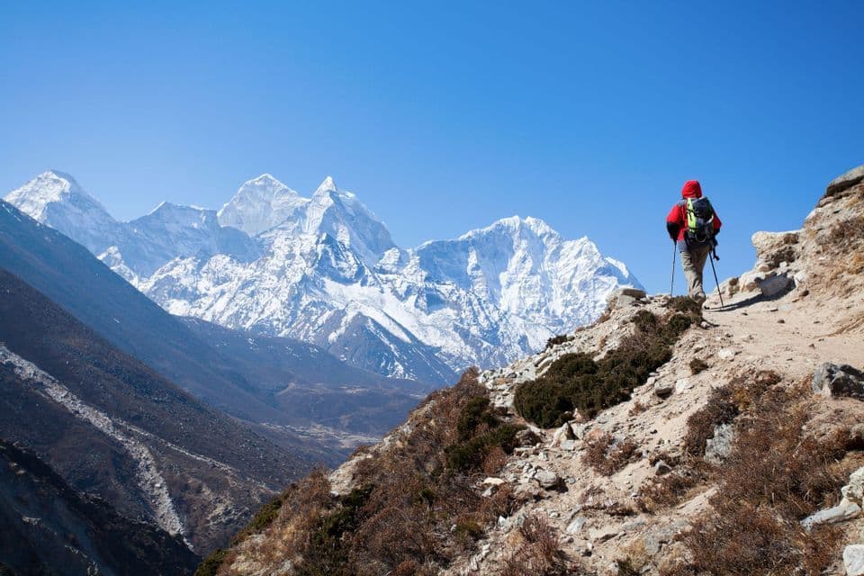 Un senderista con mochila y bastones se encuentra en un sendero rocoso, contemplando una vasta cordillera nevada bajo un cielo azul claro.