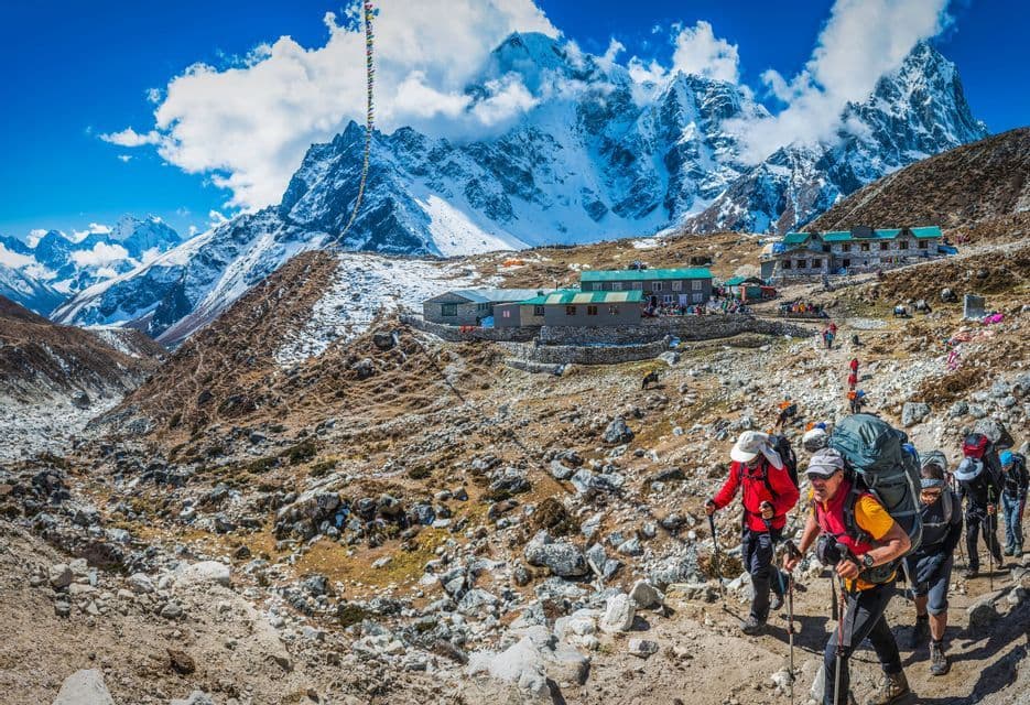 A WeRoad group trip hiking on a rocky trail towards a mountain lodge, with large snow-capped peaks in the background.
