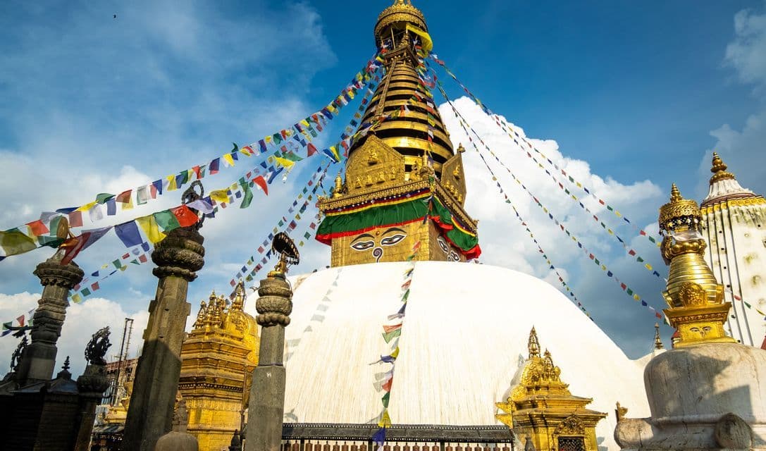 Una ripresa dal basso di un grande stupa bianco con una guglia dorata e occhi dipinti, decorato con bandiere di preghiera colorate contro un cielo blu.