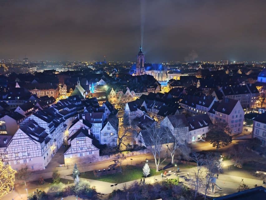 Una vista aérea de un pueblo histórico de noche, con casas de entramado de madera iluminadas y una catedral iluminada en azul con un foco.
