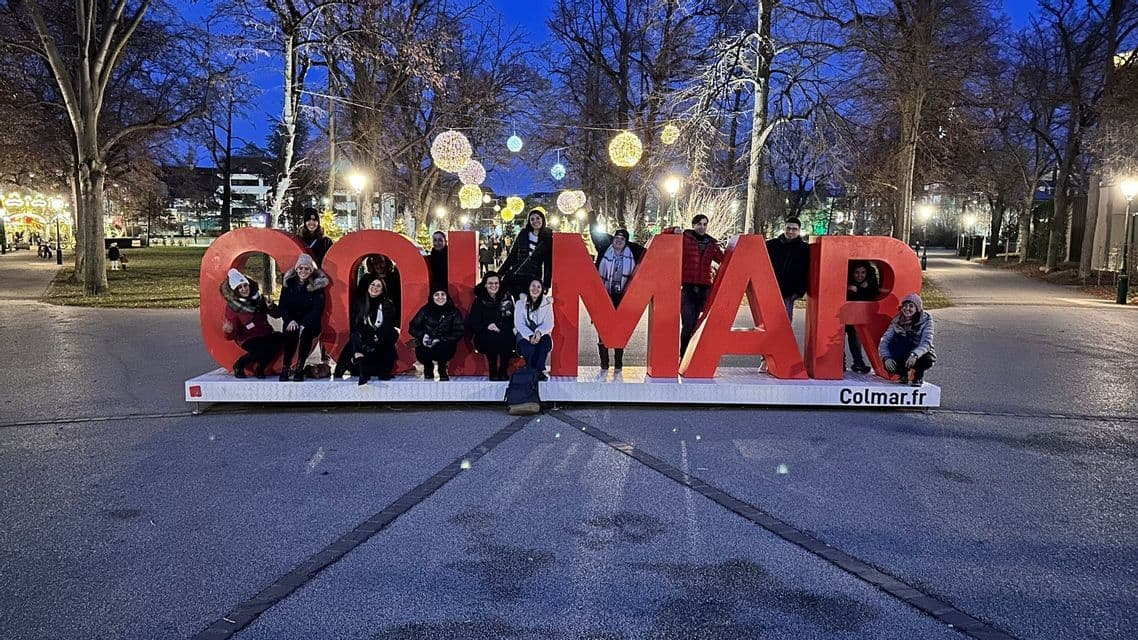 Un viaje en grupo de WeRoad posando para una foto con grandes letras rojas que forman 'COLMAR' en un parque al atardecer, con luces festivas colgando encima.