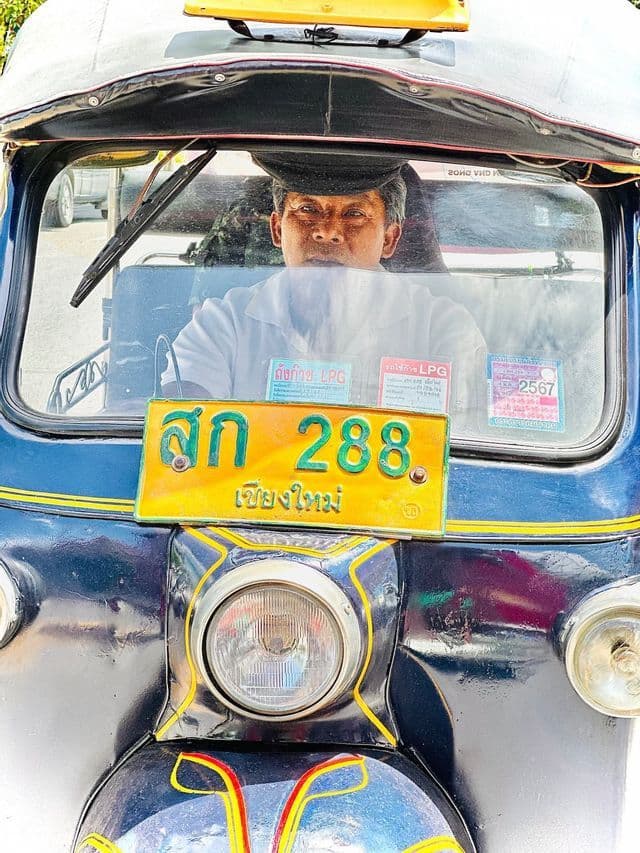A driver wearing a cap looks out from the driver's seat of a blue tuk-tuk.