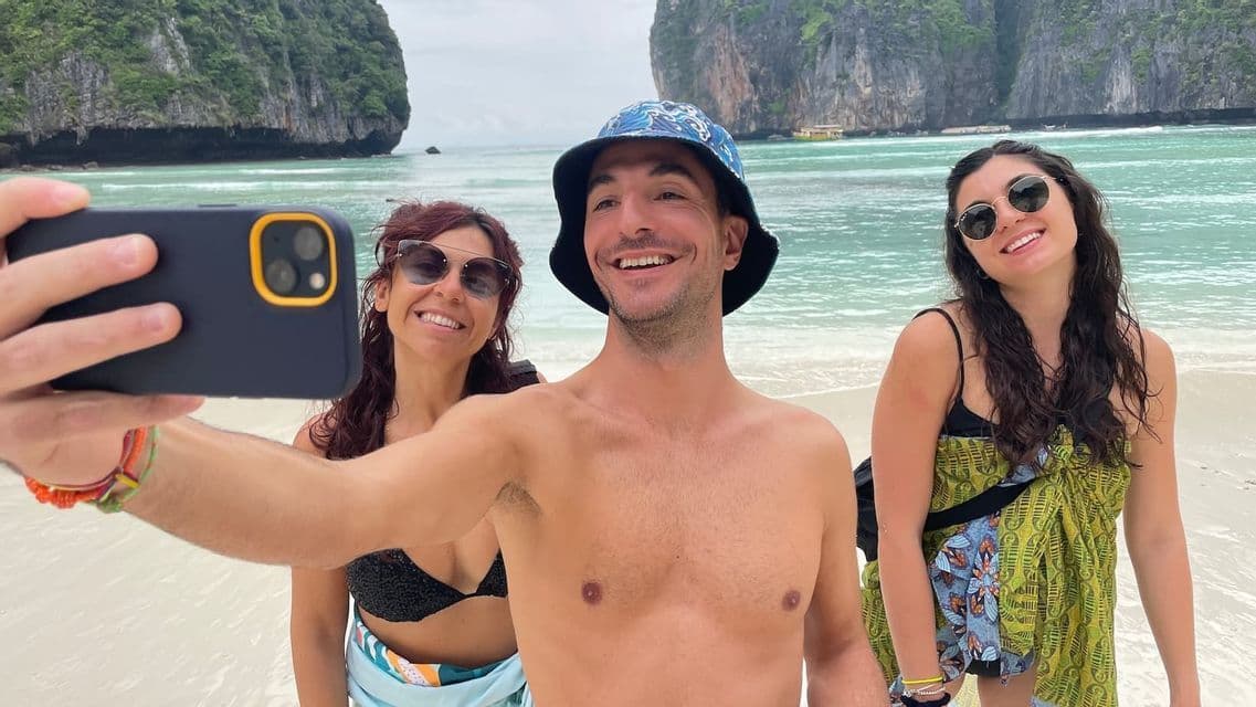 Ein Mann auf einer WeRoad Gruppenreise macht ein Selfie mit zwei lächelnden Frauen an einem Strand mit großen Klippen im Hintergrund.