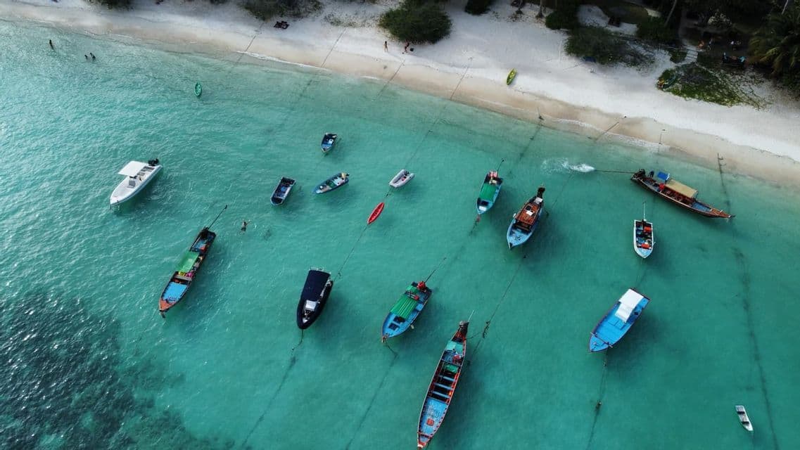 Vista aérea de coloridas embarcaciones ancladas en aguas turquesas cristalinas cerca de una playa de arena blanca.