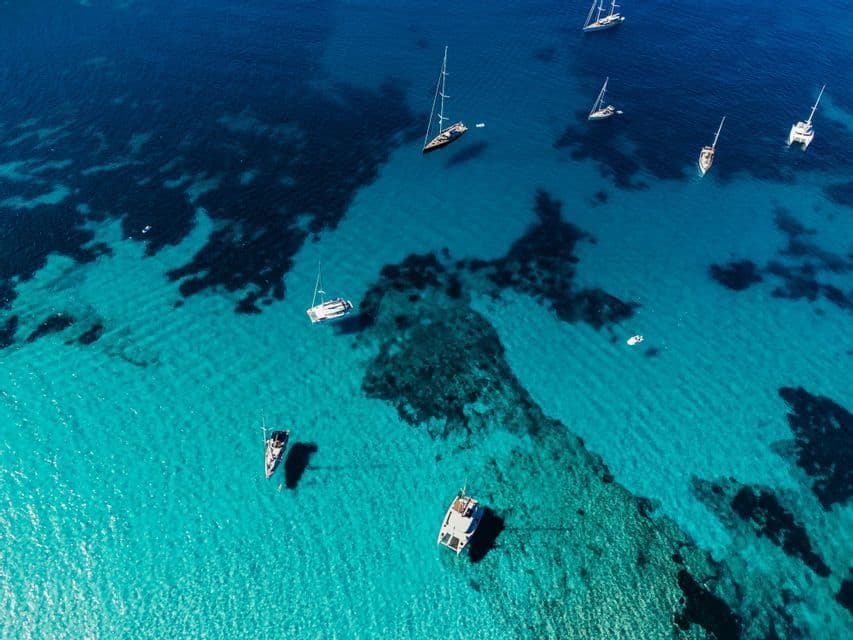 Una vista aerea di diverse barche a vela e catamarani che galleggiano in acqua trasparente turchese sopra un fondale sabbioso e roccioso.