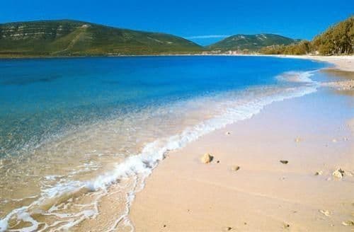 Acqua azzurra e limpida con piccole onde che accarezzano una spiaggia sabbiosa, incorniciata da verdi colline sotto un cielo azzurro brillante.