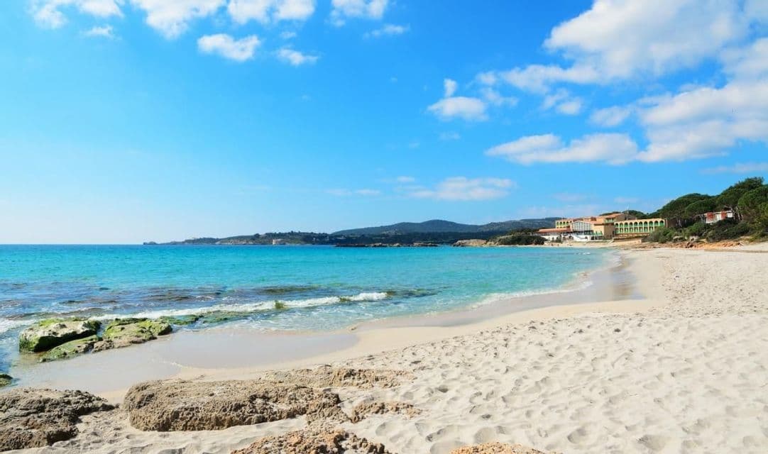 Una spiaggia di sabbia bianca incontaminata si fonde con acque cristalline turchesi, e una costa verde e collinare si staglia sullo sfondo sotto un cielo azzurro.
