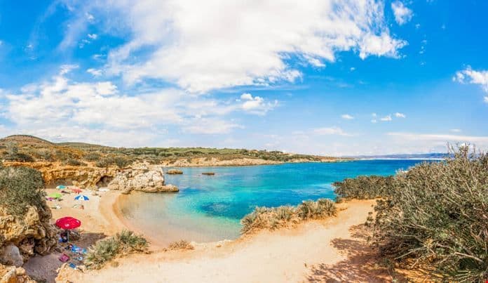 Una vista panoramica di una caletta sabbiosa appartata con acqua cristallina turchese, scogliere rocciose e ombrelloni sotto un cielo azzurro.
