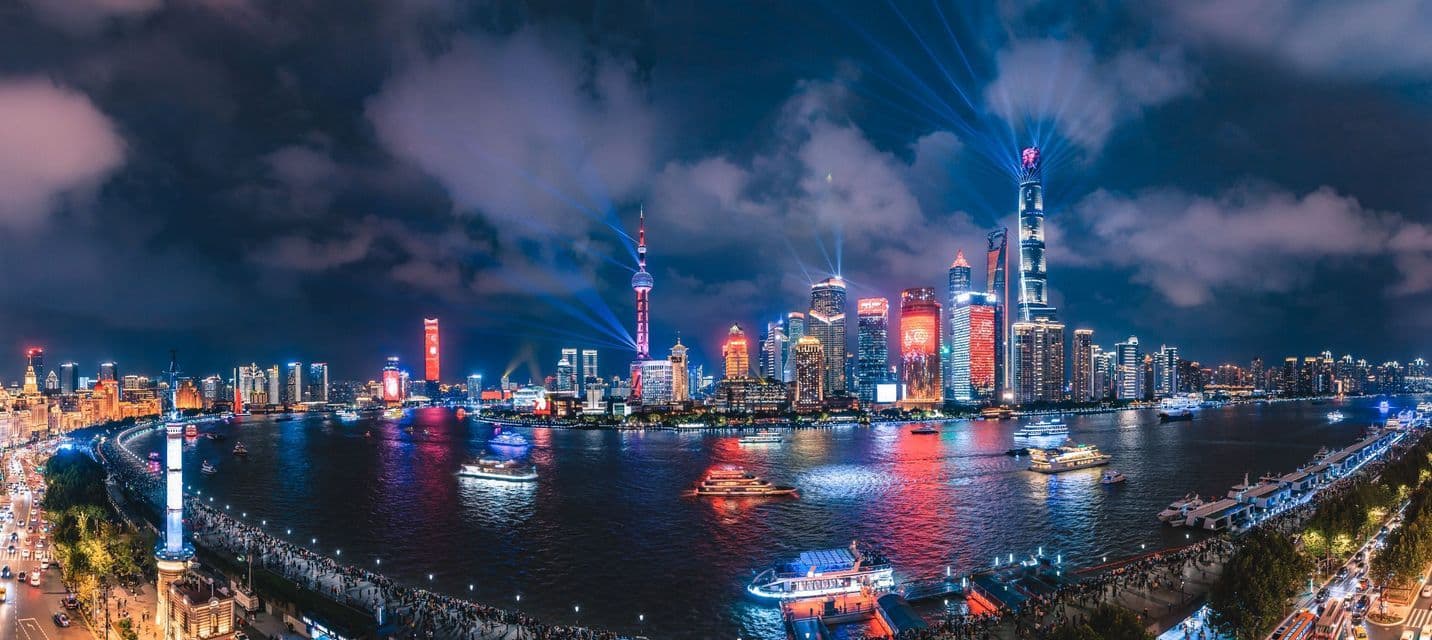 Una vista panoramica della città di notte con grattacieli illuminati, fasci di luce e imbarcazioni su un ampio fiume sotto un cielo nuvoloso