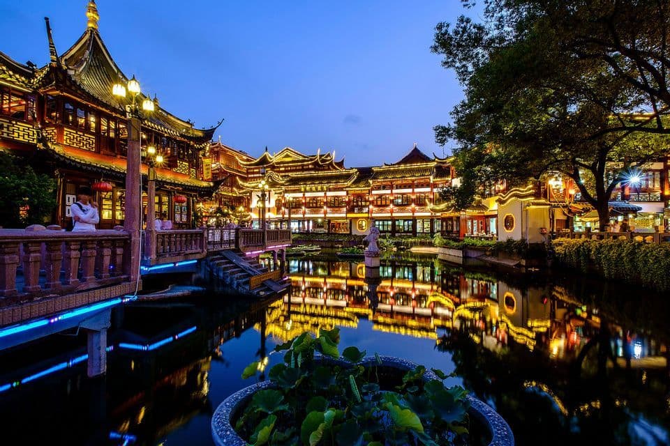 Edifici in stile tradizionale e un ponte illuminati di notte, che si riflettono in un placido stagno sotto un cielo blu scuro.