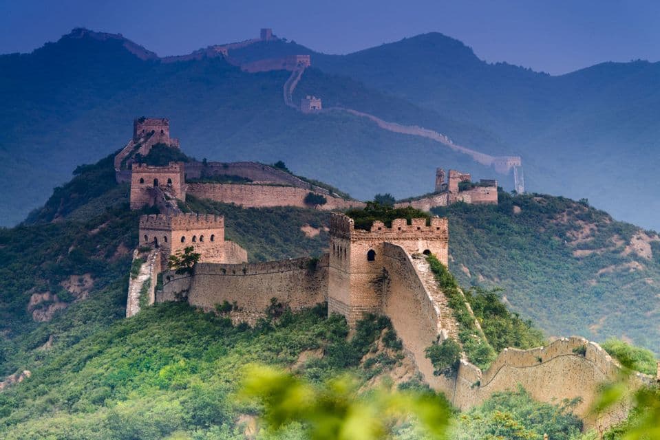 Un lungo muro di pietra con torri di guardia serpeggia sulle cime di lussureggianti montagne verdi sotto un cielo blu intenso e velato.