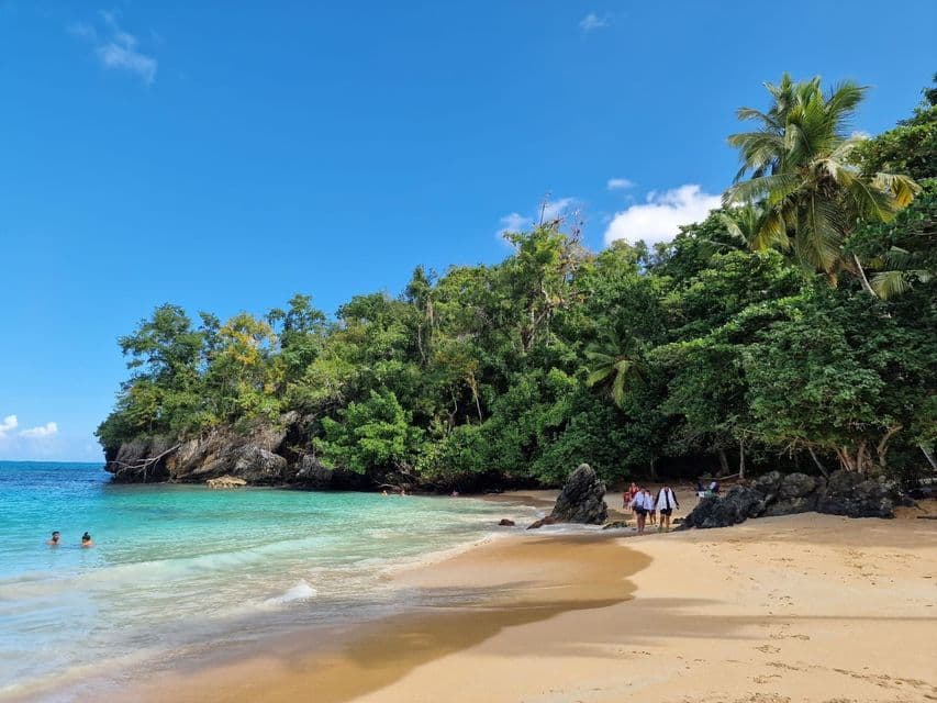 Un viaje en grupo de WeRoad camina por la orilla de una playa tropical con agua turquesa y una colina frondosa y boscosa.