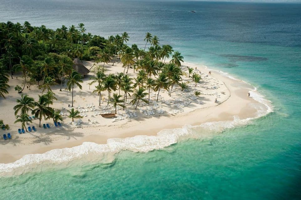 An aerial view of a white sand peninsula covered in palm trees and lined with beach loungers, surrounded by turquoise water.