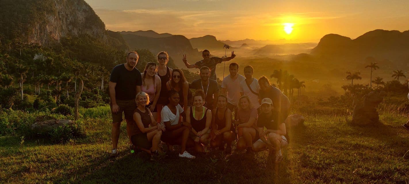 Un groupe WeRoad prend la pose pour une photo sur une colline herbeuse dominant une vallée avec des montagnes et des palmiers au coucher du soleil.