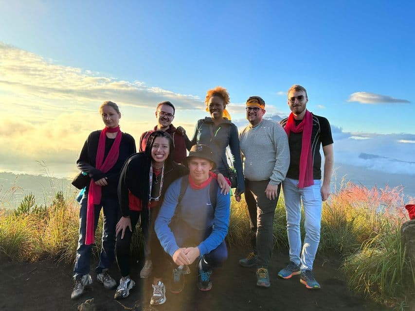 Un voyage de groupe WeRoad de sept personnes posant pour une photo au sommet d'une montagne au lever du soleil, avec une vue panoramique sur les nuages en contrebas.