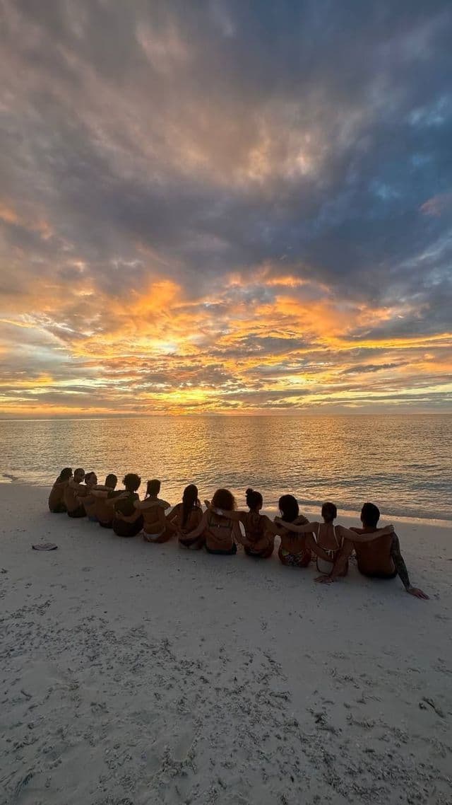 Un gruppo WeRoad seduto in fila su una spiaggia sabbiosa, ad ammirare un tramonto colorato sull'oceano.