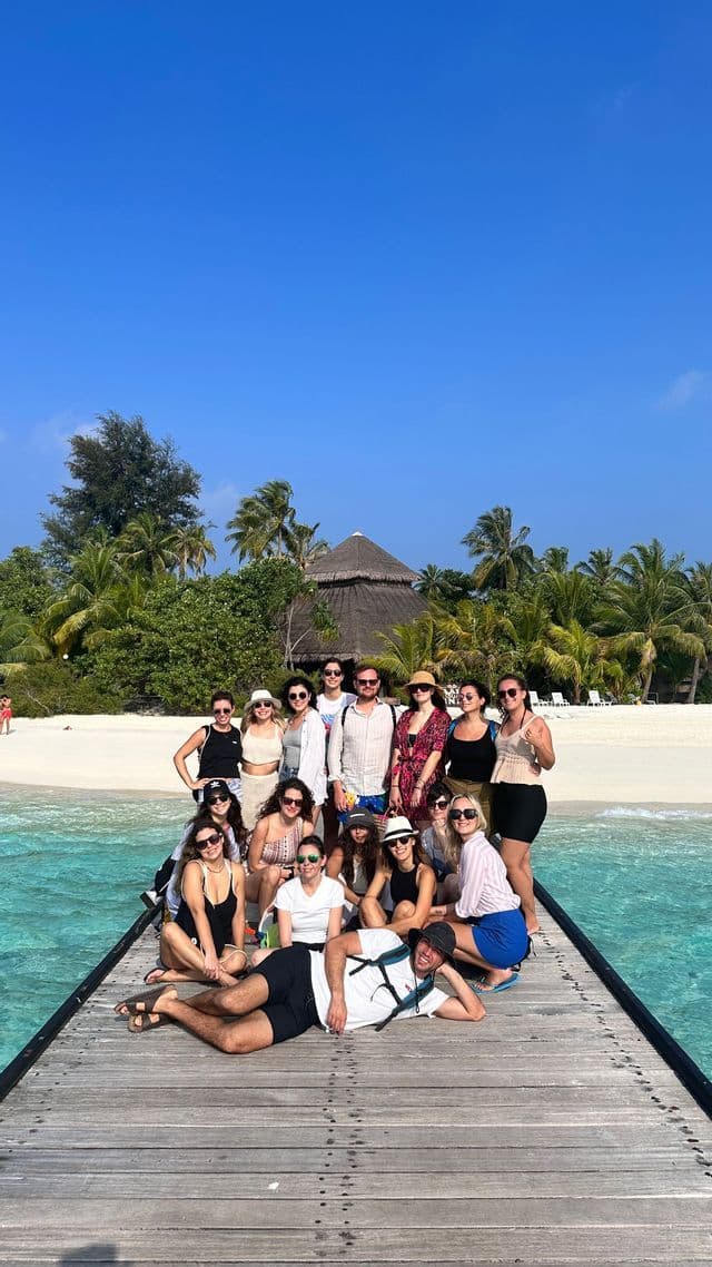 A WeRoad group trip poses for a photo on a wooden pier over turquoise water, with a tropical beach and palm trees behind them.