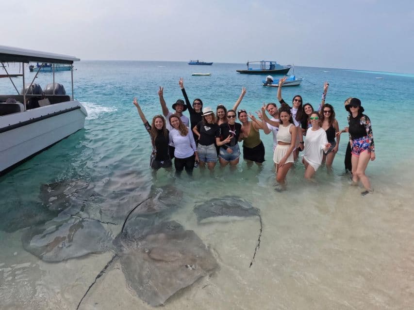 A WeRoad group trip poses in shallow, clear water on a tropical beach with several large stingrays swimming at their feet.