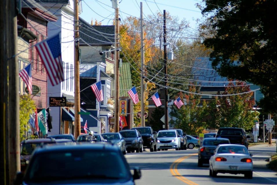 Una vista su una strada di una piccola città fiancheggiata da negozi, auto e bandiere americane sotto un limpido cielo autunnale.