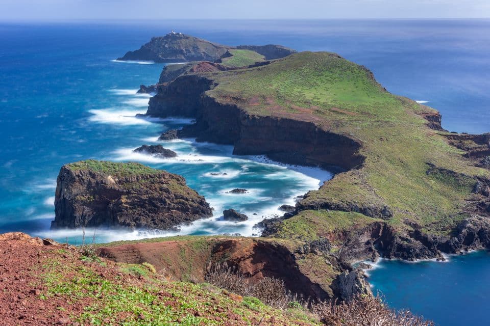 Una foto a lunga esposizione di una penisola verde e rocciosa che si protende nell'oceano blu, con onde che si infrangono sulle scogliere.