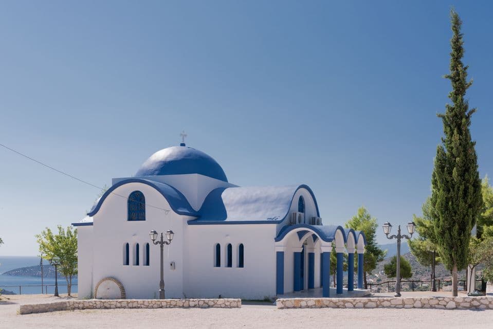 Una iglesia blanca, con una cúpula azul y detalles en el tejado, se alza en una colina con vistas al mar bajo un cielo despejado.