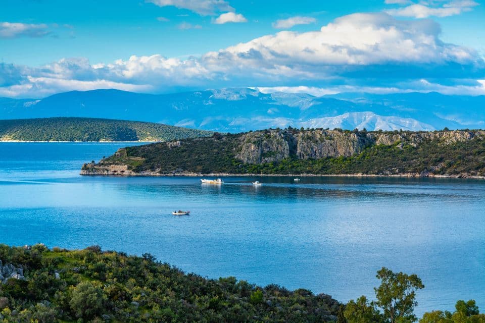 Pequeños barcos en un mar azul tranquilo con una costa rocosa verde y montañas distantes bajo un cielo parcialmente nublado.
