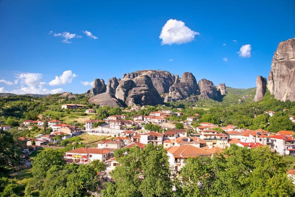 A village with red-tiled roofs is nestled among green trees at the base of towering rock formations under a blue sky.