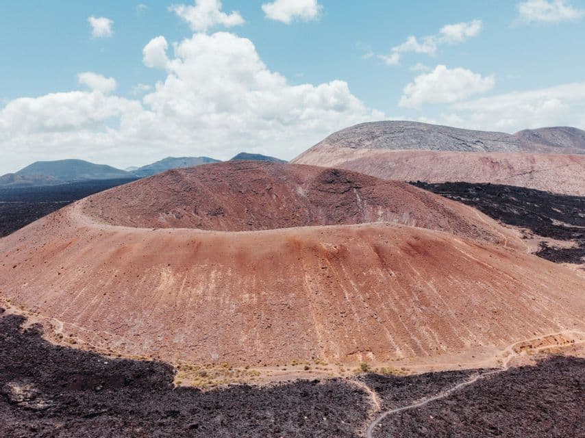 Eine Luftaufnahme eines großen, rotbraunen Vulkankraters, umgeben von schwarzen Lavafeldern, unter blauem Himmel mit weißen Wolken.