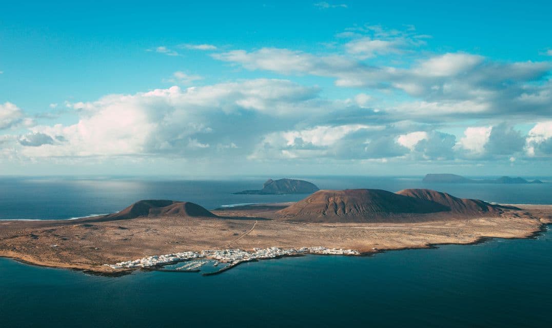 Vue aérienne d'une île volcanique avec un petit village côtier et un port, entourée d'un océan bleu profond sous un ciel partiellement nuageux.