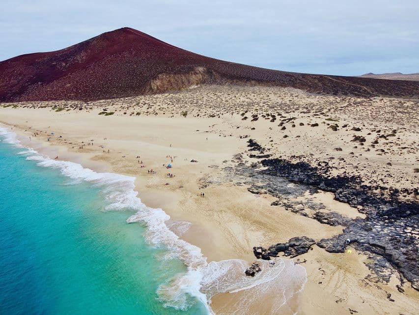 Vue aérienne d'une plage de sable avec de l'eau turquoise et des roches volcaniques, avec une grande montagne rouge en arrière-plan.