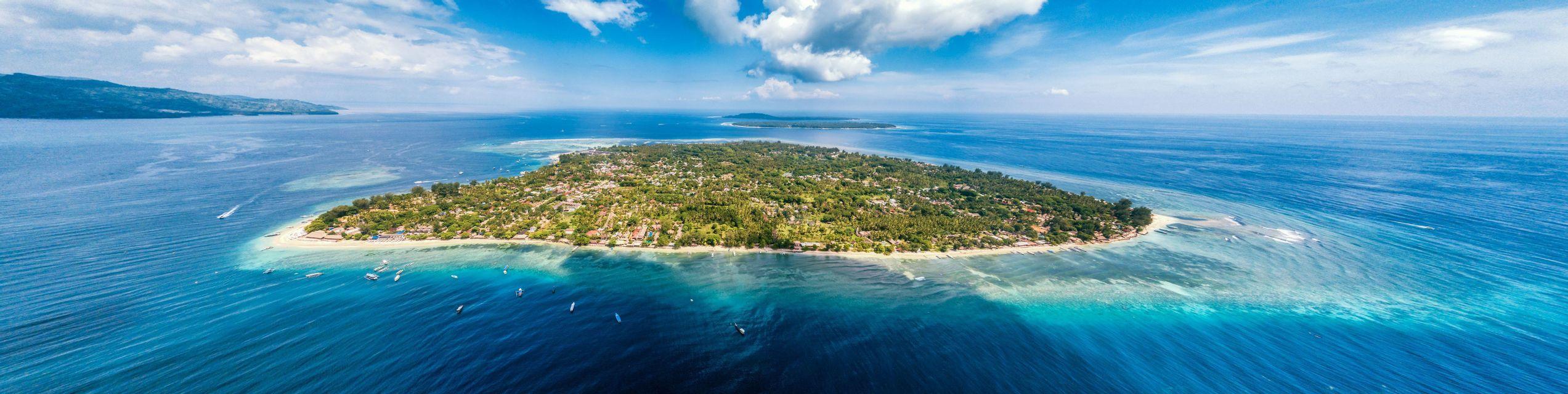 Eine Panorama-Luftaufnahme einer üppigen Tropeninsel, umgeben von türkisfarbenem Ozean, mit verstreuten Booten auf dem Wasser unter bewölktem Himmel.