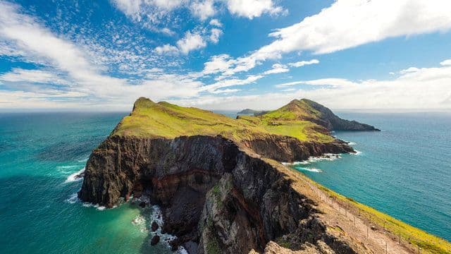 Una penisola rocciosa coperta da erba verde si protende in un oceano turchese, con un sentiero lungo la sua cresta sotto un cielo blu.