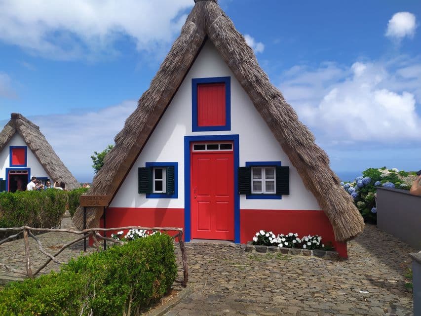 Una tradizionale casa a forma di A con tetto di paglia e vivaci finiture rosse e blu si erge su un sentiero acciottolato sotto un cielo azzurro.