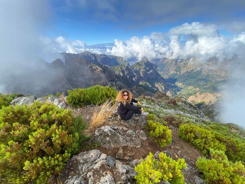 Una donna con capelli ricci e occhiali da sole siede su una cima rocciosa di montagna che domina una valle panoramica con nuvole sottostanti.